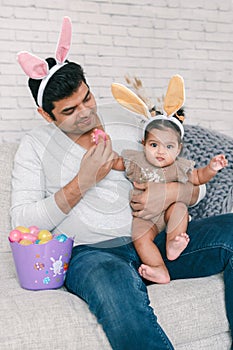 Indian father with baby girl daughter in bunny ears playing toy eggs in basket celebrating Easter