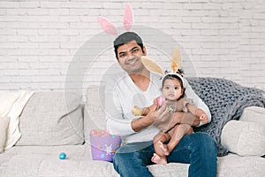 Indian father with baby girl daughter in bunny ears playing toy eggs in basket celebrating Easter