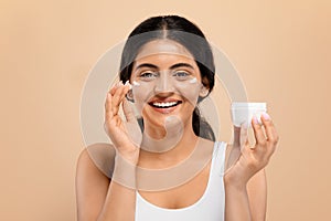 Indian Female Applying Moisturising Cream On Face While Standing Against Beige Background