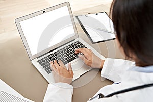 Indian female doctor typing using laptop mock up white screen at desk, close up.