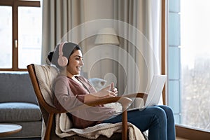Indian female in headphones sit in comfy chair use laptop