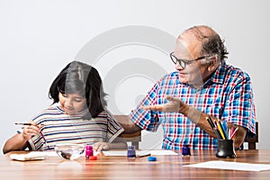 Indian grandfather helping grand daughter drawing & painting with colours and brush on paper at home