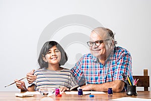 Indian grandfather helping grand daughter drawing & painting with colours and brush on paper at home