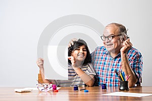 Indian grandfather helping grand daughter drawing & painting with colours and brush on paper at home