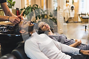 Indian male client having his hair washed by hairdresser next to happy blonde female customer in a professional hair