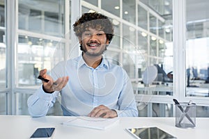 Indian male spreading hands with pen to camera while sitting in front of table with notebook. Smiling hr manager asking