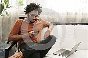 Indian male using his phone, laptop at arm's reach, in a warm room setting