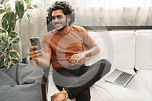 Indian male using his phone, laptop at arm& x27;s reach, in a warm room setting