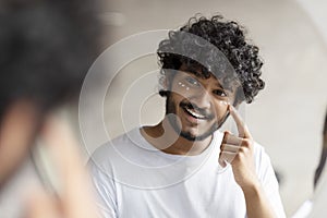 Indian man applying cream under her eyes and smiling at his reflection in mirror, enjoying morning beauty routine