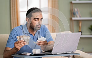 Indian man calculating monthly expenses from bills after salary while sitting on sofa at home - concept of Budget