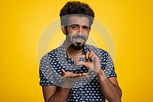 Indian man showing a little bit gesture with sceptic smile showing minimum sign measuring small size