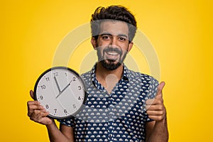 Indian man showing time on wall office clock, ok, thumb up, approve, pointing finger at camera