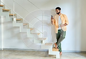 Indian man wearing earbud using smartphone standing at home.