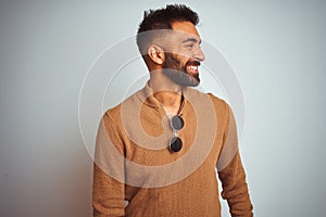 Indian man wearing elegant sweater and sunglasses standing over isolated white background looking away to side with smile on face,