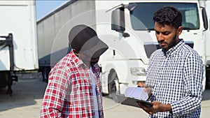 Indian manager and african trucker standing by truck with a clipboard, checking the delivery list. Two workers preparing