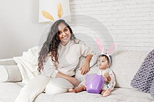 Indian mother with baby girl daughter in bunny ears playing toy eggs in basket celebrating Easter