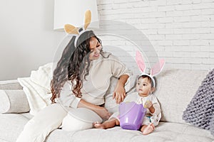 Indian mother with baby girl daughter in bunny ears playing toy eggs in basket celebrating Easter