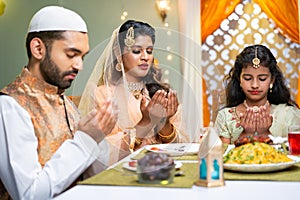 Indian Muslim family with kid praying at ramadan iftar dinner before eating at home - concept of traditional festival