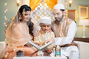 Indian muslim parents helping kid to reading quran before fasting food during ramadan festival at home - concept of