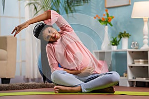 Indian pregnant woman at home doing yoga asana or exercise by sitting on yoga mat at home - concept of prenatal fitness