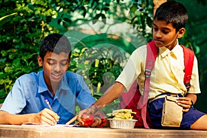 Indian school boy writing on notepad doing homework , looking concentration . His friend with backpack sits on table and