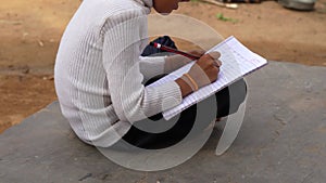 Indian schoolgirl is sitting on the ground near goat farm and writing in a book. A little girl is doing her homework in nature