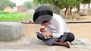 Indian schoolgirl is sitting on the ground near goat farm and writing in a book. A little girl is doing her homework in nature