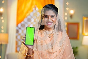 Indian smiling muslim woman showing green screen mobile phone by looking at camera during ramadan festival at home -