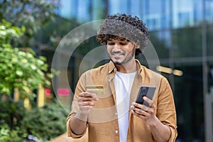 Indian smiling young man standing on city street, holding phone, looking and using credit card