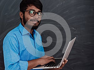 Indian smiling young student in blue shirt and glasses using laptop and posing on school blackboard background
