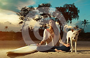 Indian surfer girl meditating in lotus pose on the beach at sunset next to surfboard