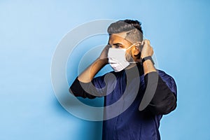 Indian surgeon standing setting his gloves to start the surgery, wearing a mask and a blue uniform, isolated on a blue background