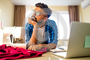 Indian tailor asian man designer sewing clothes on sewing machine next to laptop home apartment workplace