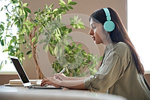 Indian teen girl student wear headphones watching online class making notes.