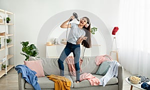 Indian teen girl wearing headphones, using blowdryer as microphone, singing song and dancing on sofa at home