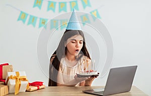 Indian teenage girl in festive hat having online birthday party, blowing candles on cake in front of laptop webcam