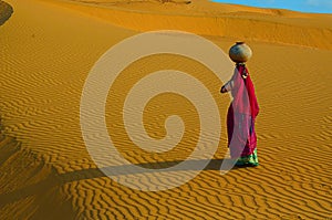 Indian woman carrying heavy jug of water on her head and walking on a yellow sand dune in the hot summer desert against blue sky.