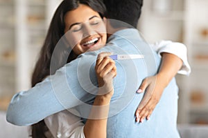 Indian woman joyfully hugging man, holding positive pregnancy test