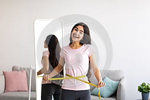 Indian woman measuring her waist with tape in front of mirror, showing results of slimming diet or liposuction at home