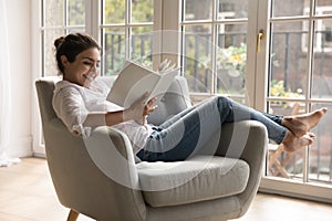 Indian woman reading book seated on cozy armchair