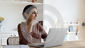 Indian woman sit at table with laptop thinking looks aside