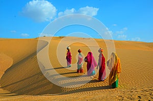 Indian women carrying heavy jugs of water on their head and walking on a yellow sand dune in the hot summer desert against blue