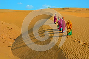 Indian women carrying heavy jugs of water on their head and walking on a yellow sand dune in the hot summer desert against blue