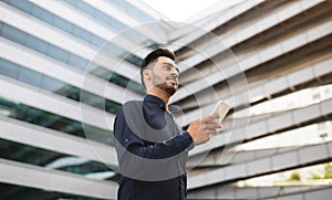 Indian young businessman posing with smartphone against modern office building