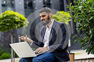 Indian young male businessman sitting on a bench near an office center wearing a suit and headphones, holding a laptop