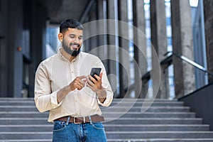 Indian young male student standing on campus on stairs near building wearing shirt and jeans and smilingly using mobile photo