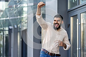 Indian young man standing near an office building and rejoicing in success raising his hand up and shouting with