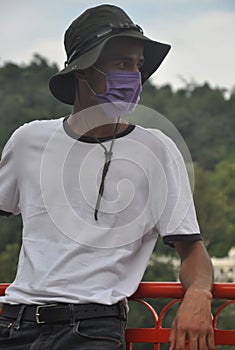 A Indian young man standing outside with wearing boonie hat, white t-shirt and face mask during coronavirus pandemic with looking