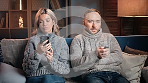 Indifferent married couple sitting couch chatting smartphone watching TV separated at evening home