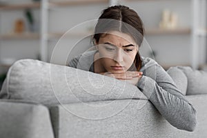 Indifferent upset frustrated european young female sits on sofa looking at emptiness at home